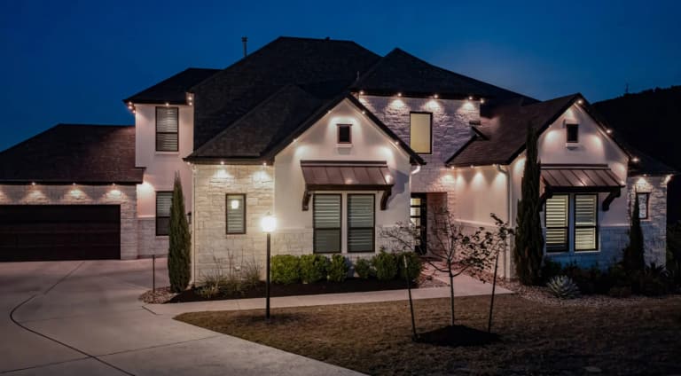 Stone and stucco home at dusk with warm white permanent lights along the eaves washing the walls below